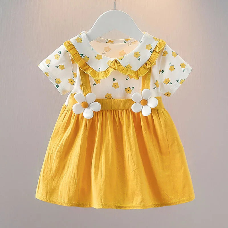Yellow girl's dress with flower prints and white floral decorations on the straps. Features a ruffled collar and short sleeves with matching floral patterns on a white base.