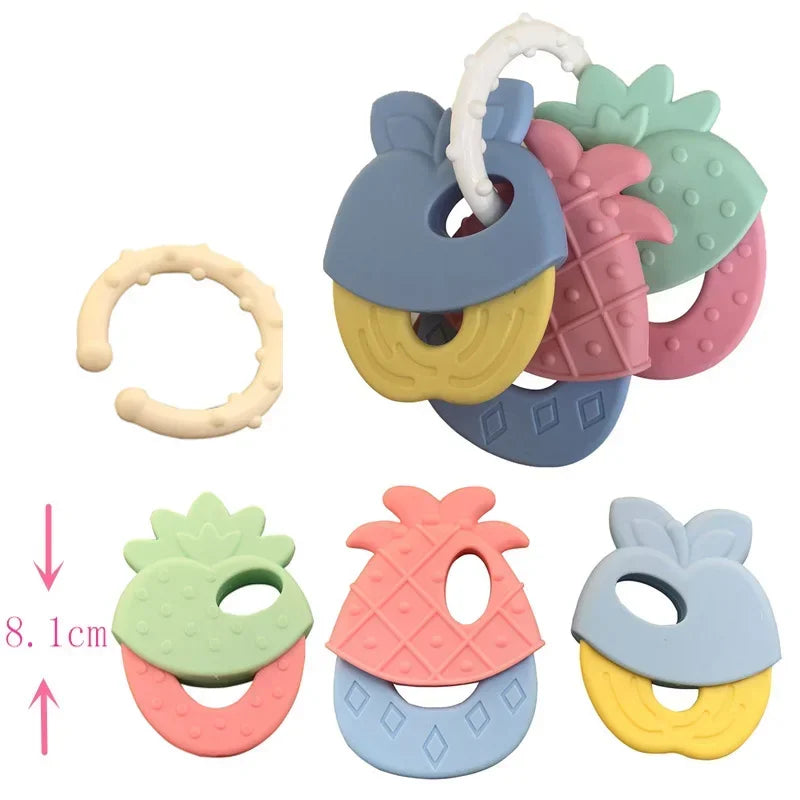 Colorful fruit-shaped baby teethers in pastel shades, including pineapple, strawberry, and blueberry designs. Teethers are linked by a ring, with a single detachable ring shown.