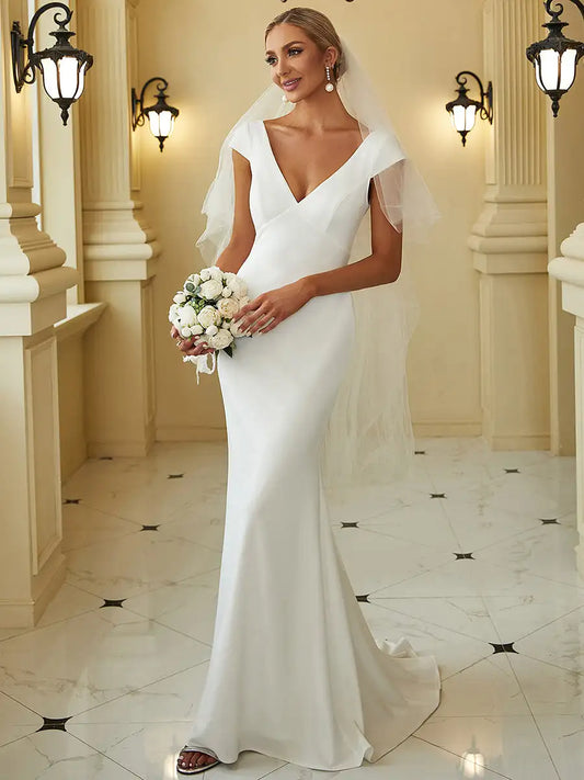 Bride in an elegant white gown with a deep V-neck and veil, holding a bouquet of white flowers. She stands in a classic hallway with ornate wall sconces and a tiled floor.