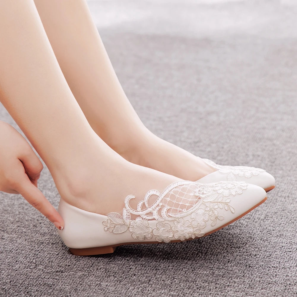 Person wearing elegant white flats with floral lace embroidery, pointing at the heel while sitting on a soft gray carpet.