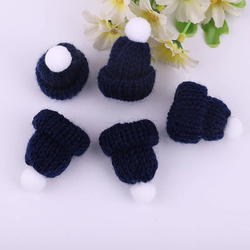 Five miniature navy blue knit hats with white pom-poms arranged on a white surface near pale flowers with yellow centers.
