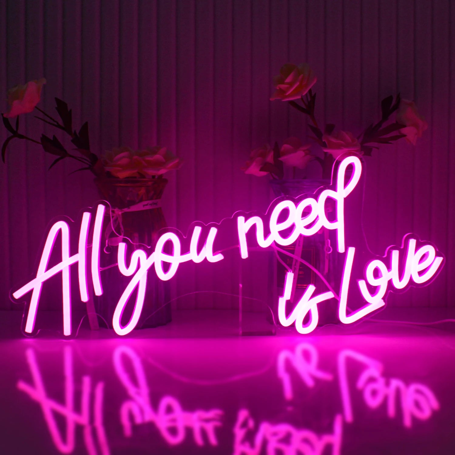Pink neon sign saying "All you need is Love" with a reflection on a glossy surface and roses in the background against a striped wall.