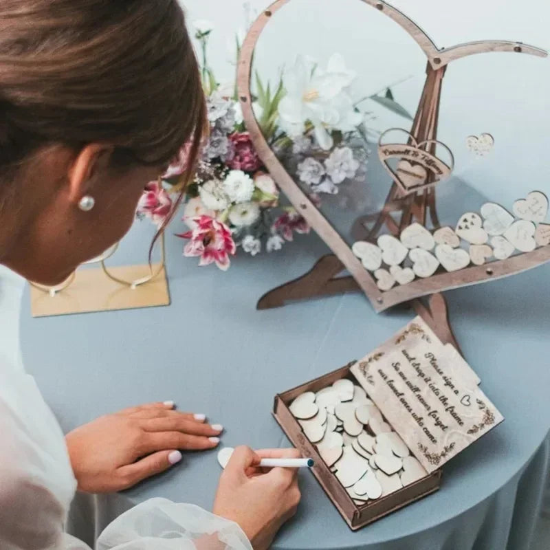 Woman writing on a heart-shaped wooden piece at a wedding guestbook table, featuring a heart frame filled with similar pieces, a bouquet of flowers, and a decorated message box.