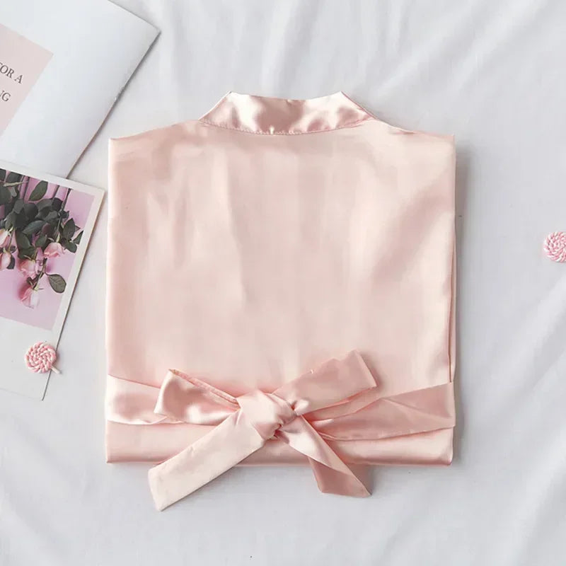 Folded blush pink silk robe with a wide belt tied in a bow, placed on a white surface. Nearby are a floral magazine page and pink decorative elements.