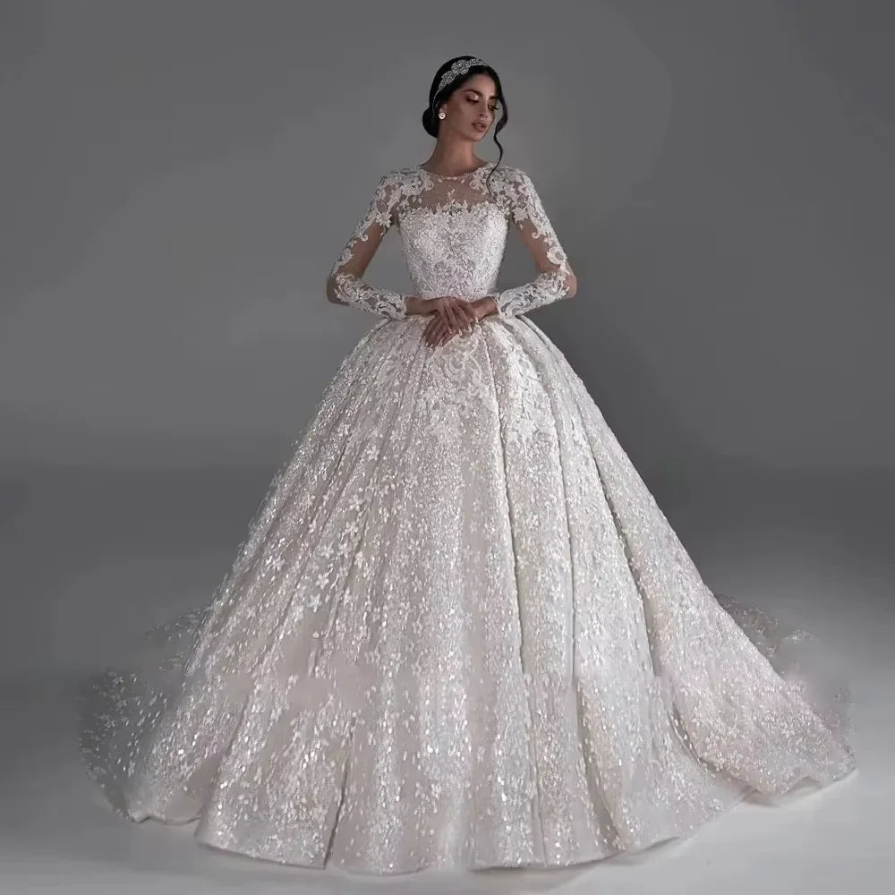 Elegant bride in a sparkling ivory ball gown with intricate lace and long sleeves, standing against a gray background; her hair is styled with a jeweled headpiece.