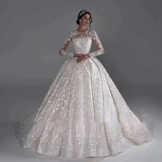 Elegant bride in a sparkling ivory ball gown with intricate lace and long sleeves, standing against a gray background; her hair is styled with a jeweled headpiece.