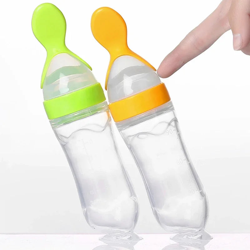 Two baby bottles with soft silicone spoon attachments, one green and one orange, with a finger lightly pressing the orange-tipped one. They are standing upright on a reflective surface.