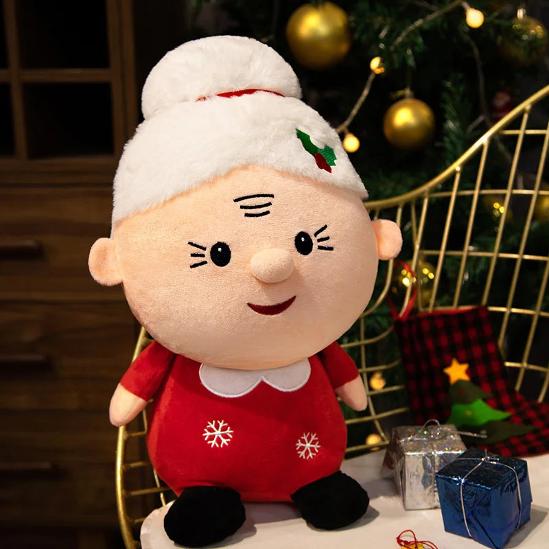A plush toy resembling Mrs. Claus with a white hat, red dress, and snowflake patterns sits on a chair. Christmas tree and wrapped gifts are in the background.