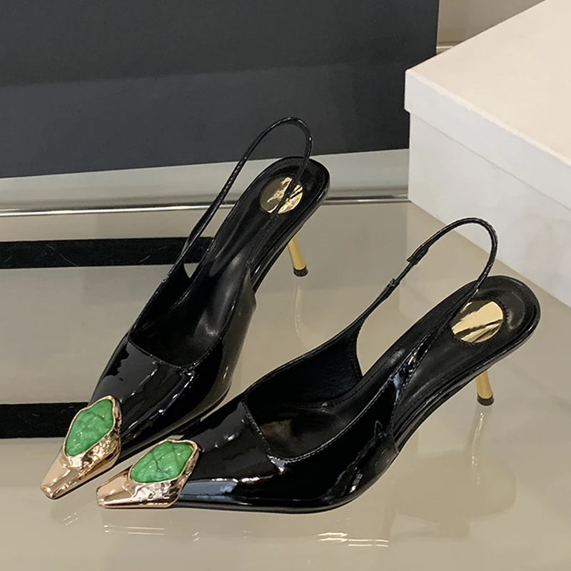 Black slingback heels with gold-tipped pointed toes, featuring a green gemstone detail. They have slender gold stiletto heels and are displayed on a reflective surface.