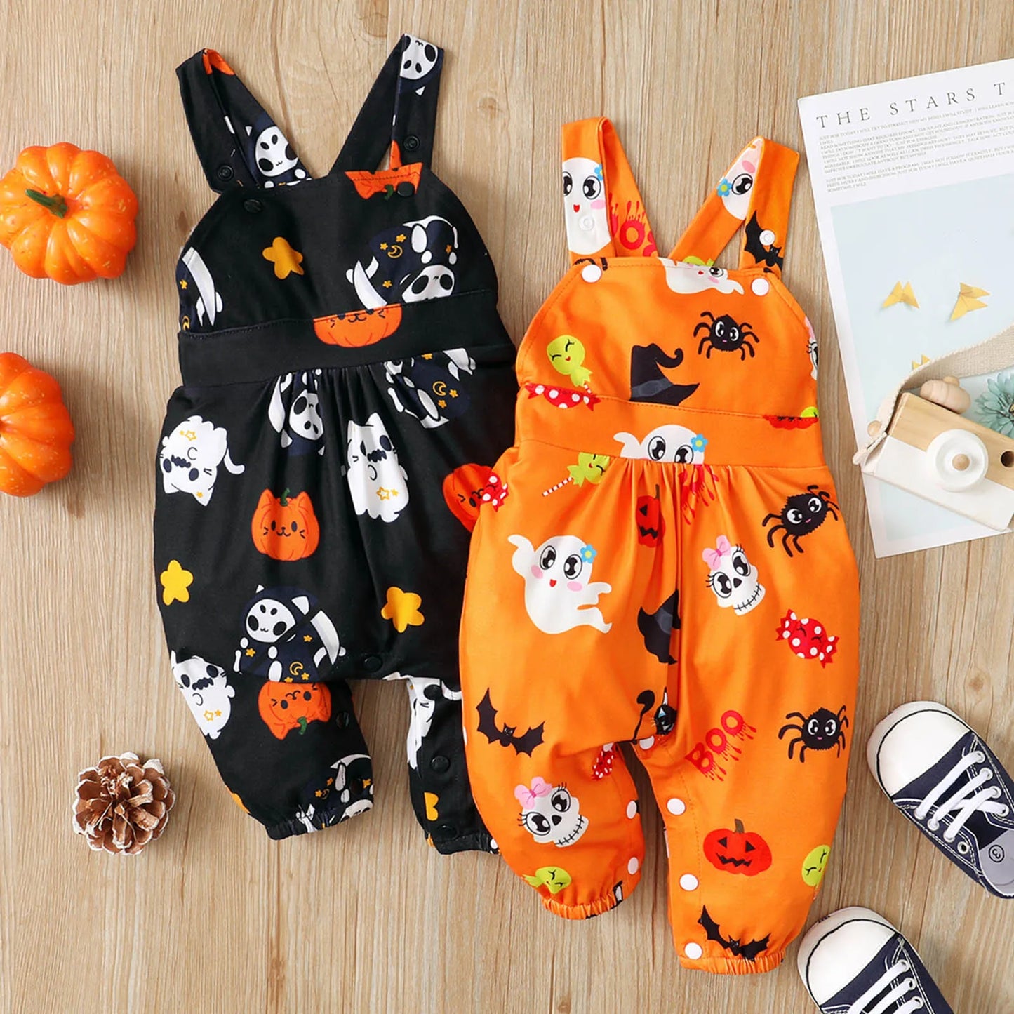 Two cute Halloween-themed baby rompers on a wooden surface, one black and one orange, decorated with ghosts, pumpkins, and bats. Nearby are small pumpkins and baby shoes.