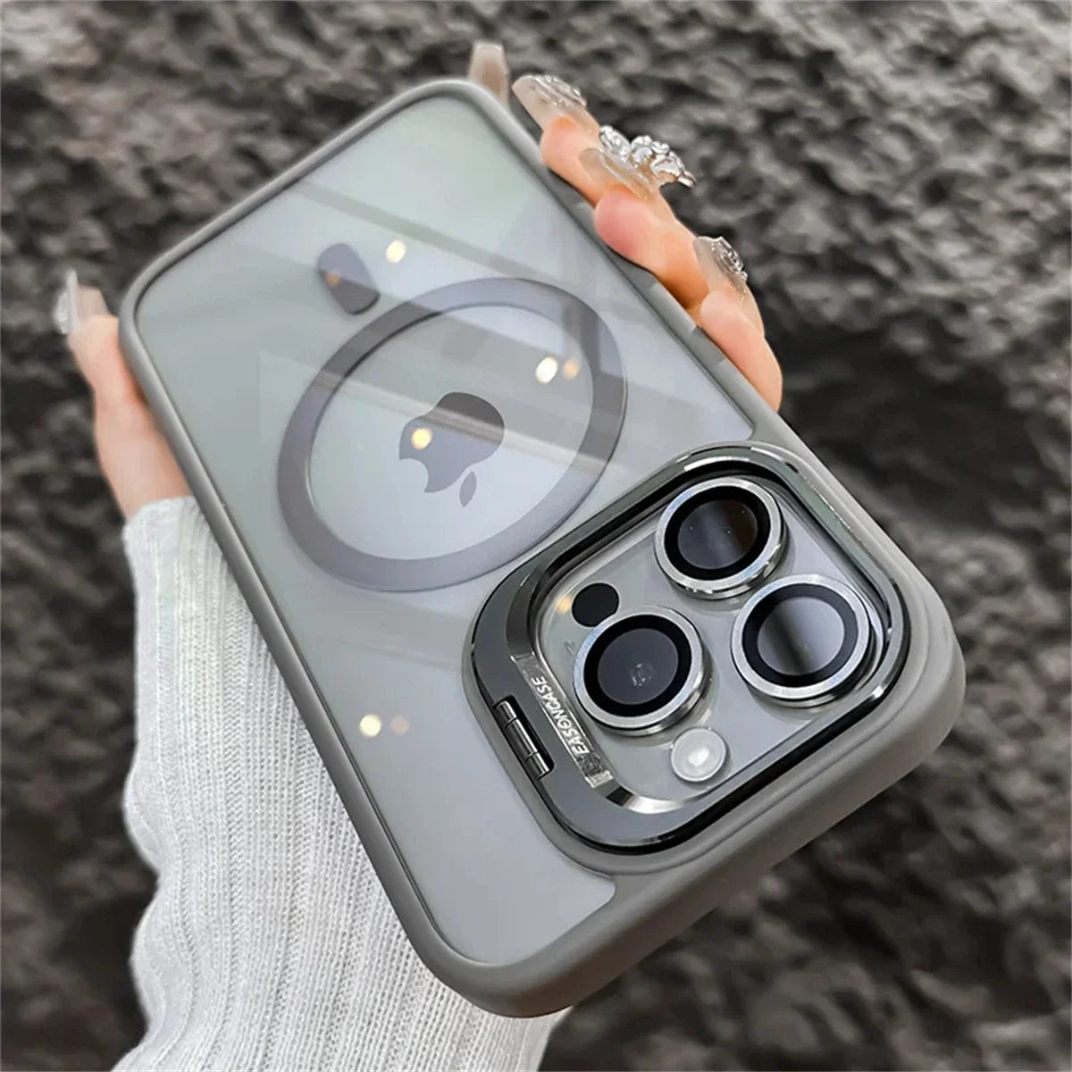 Close-up of a hand holding a smartphone with a frosted case featuring a visible Apple logo and MagSafe ring. The phone has three camera lenses and a textured, rocky background.