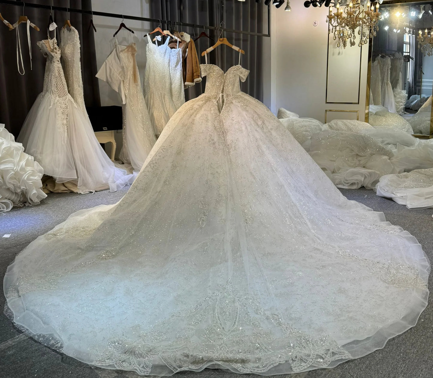 Elegant bridal boutique featuring an ornate, off-shoulder wedding gown with a long train, surrounded by various delicate dresses on hangers and showcased under a chandelier.