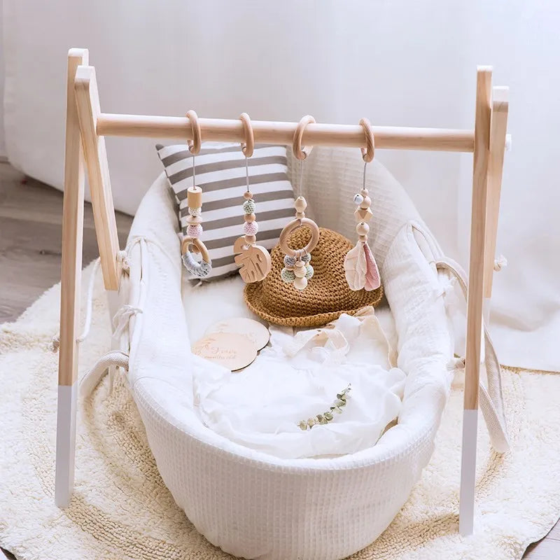 A cozy white bassinet with a striped cushion, adorned with a wooden play gym featuring pastel-colored hanging toys and a knitted brown hat. A soft rug lies underneath.