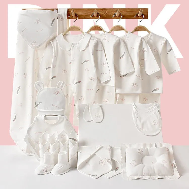 A collection of soft white baby clothing and accessories with subtle patterns, including robes, shirts, bibs, socks, and a pillow, displayed on wooden hangers against a pink background.