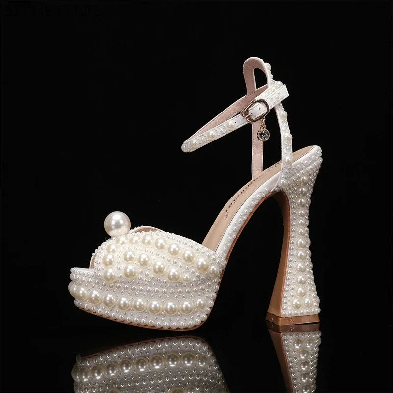 Elegant high-heeled shoe adorned with pearls, featuring an ankle strap and a large pearl accent on the toe. The shoe has a glossy finish and a square heel.