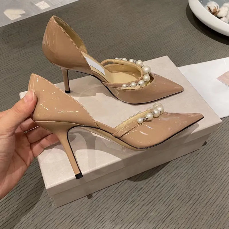 Beige patent leather high heels with pearl adornments on the strap are being held above a matching shoe box, placed on a textured grey surface.