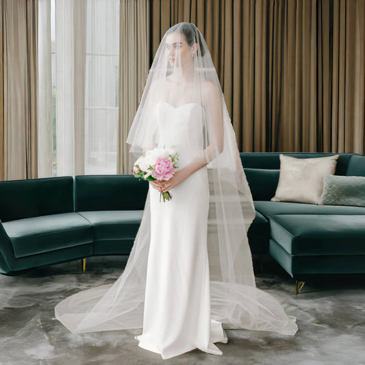 A bride in a white, strapless gown with a long veil holds a bouquet of pink and white flowers. She stands in a modern room with a dark green sofa and floor-to-ceiling curtains.