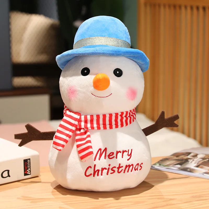 Plush snowman with a blue hat, orange nose, striped red scarf, and embroidered "Merry Christmas." It has twig-like arms and rosy cheeks, sitting on a wooden surface.