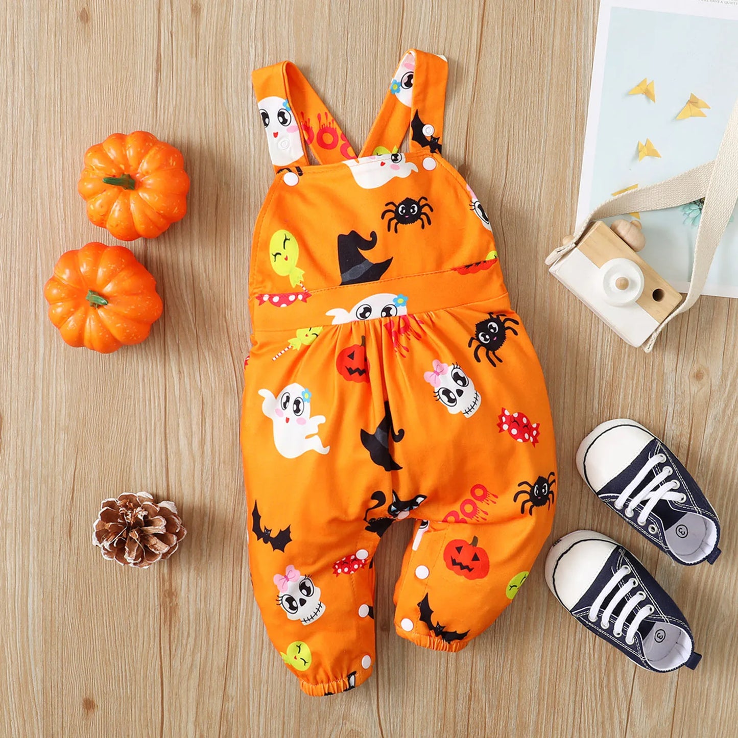 Orange Halloween-themed toddler overalls with ghost, bat, and pumpkin patterns, arranged with baby sneakers, a toy camera, two small pumpkins, and a pinecone on wooden floor.