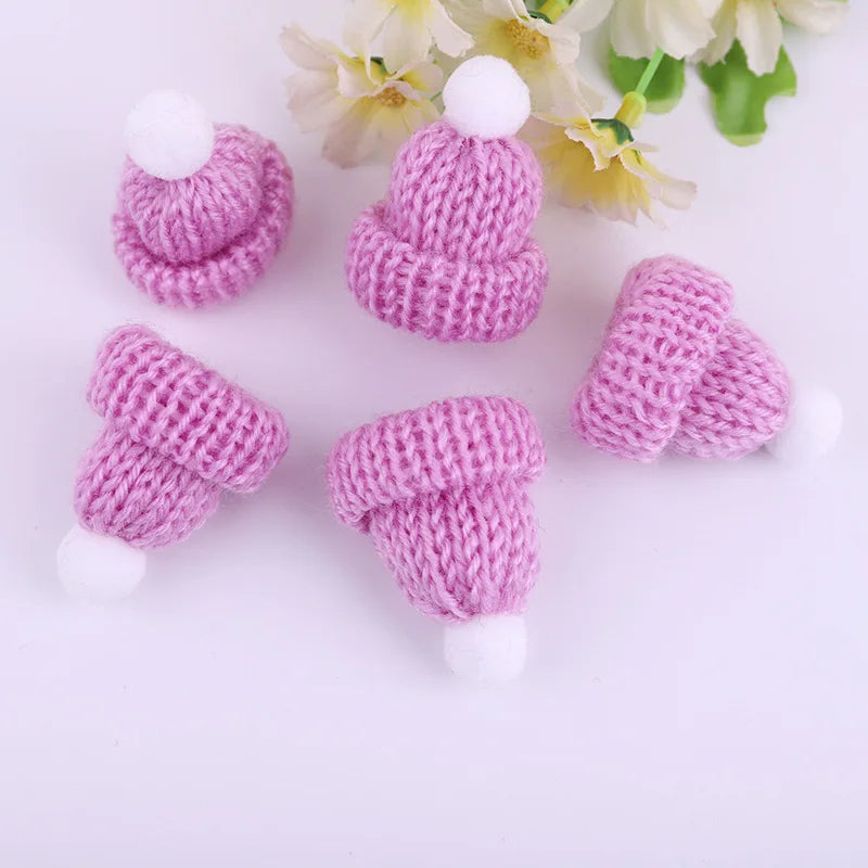 Five pink knitted mini hats with white pom-poms are arranged on a white surface. Yellow and white flowers with green leaves are visible in the background.