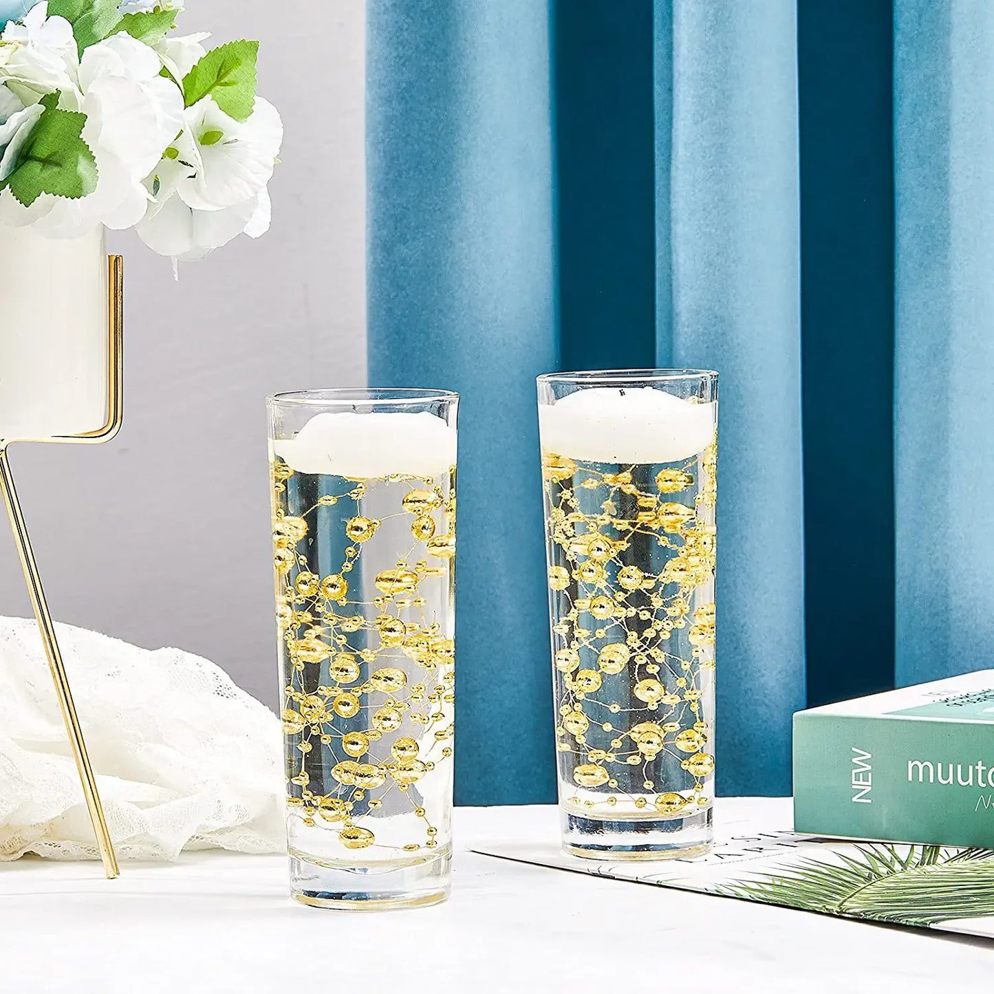 Two tall glasses with a bubbly beverage and gold bead decorations. A potted plant, blue curtains, and a green book titled "muuto" are in the background.