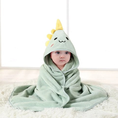 A baby wrapped in a soft green dinosaur-themed hooded towel with a smiling face and yellow spikes sits on the floor. The background is a simple, light-colored room.