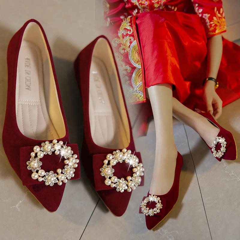 Red velvet flats adorned with pearl and crystal brooches. A person in red embroidered attire is wearing one shoe, showcasing a luxurious and elegant fashion style.