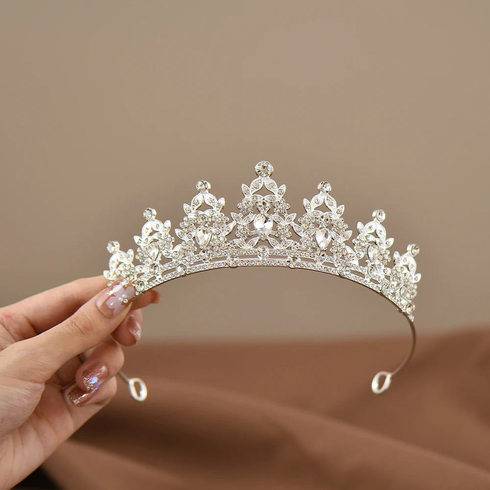 A hand holds an ornate silver tiara adorned with shimmering gemstones. The tiara features intricate floral and teardrop-shaped designs, set against a soft brown background.