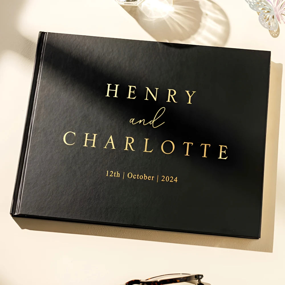 Black hardcover book with gold embossed text "Henry and Charlotte" and date "12th October 2024." Shadows and a decorative butterfly detail are visible.