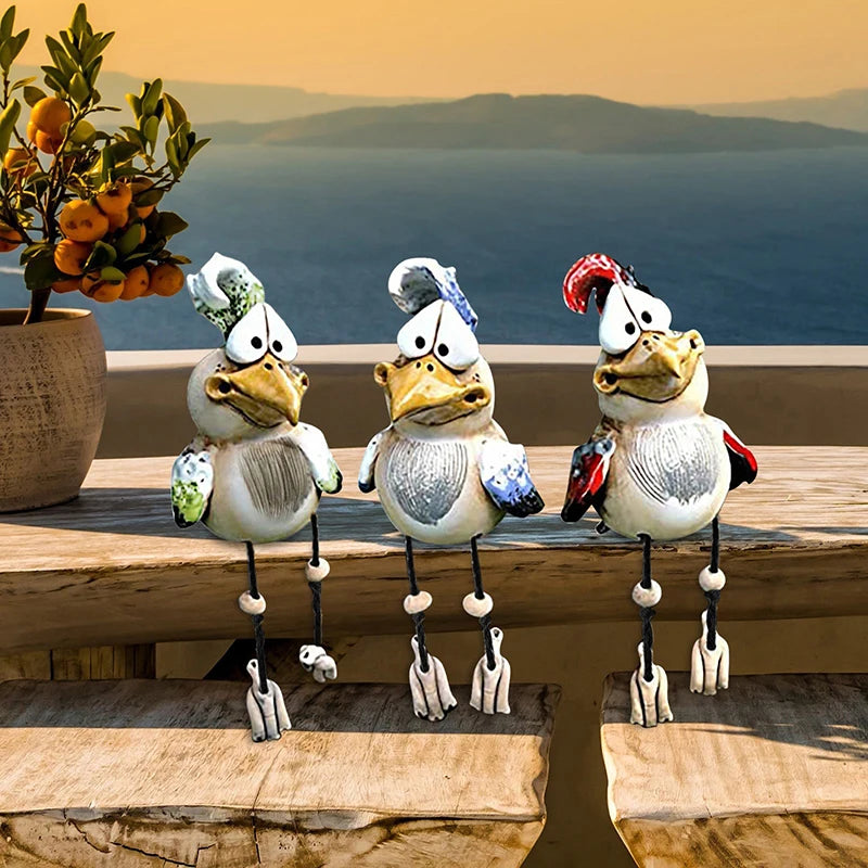 Three quirky bird figurines with colorful wings and legs sit on a wooden ledge, with a potted plant beside them and a serene seascape in the background at sunset.