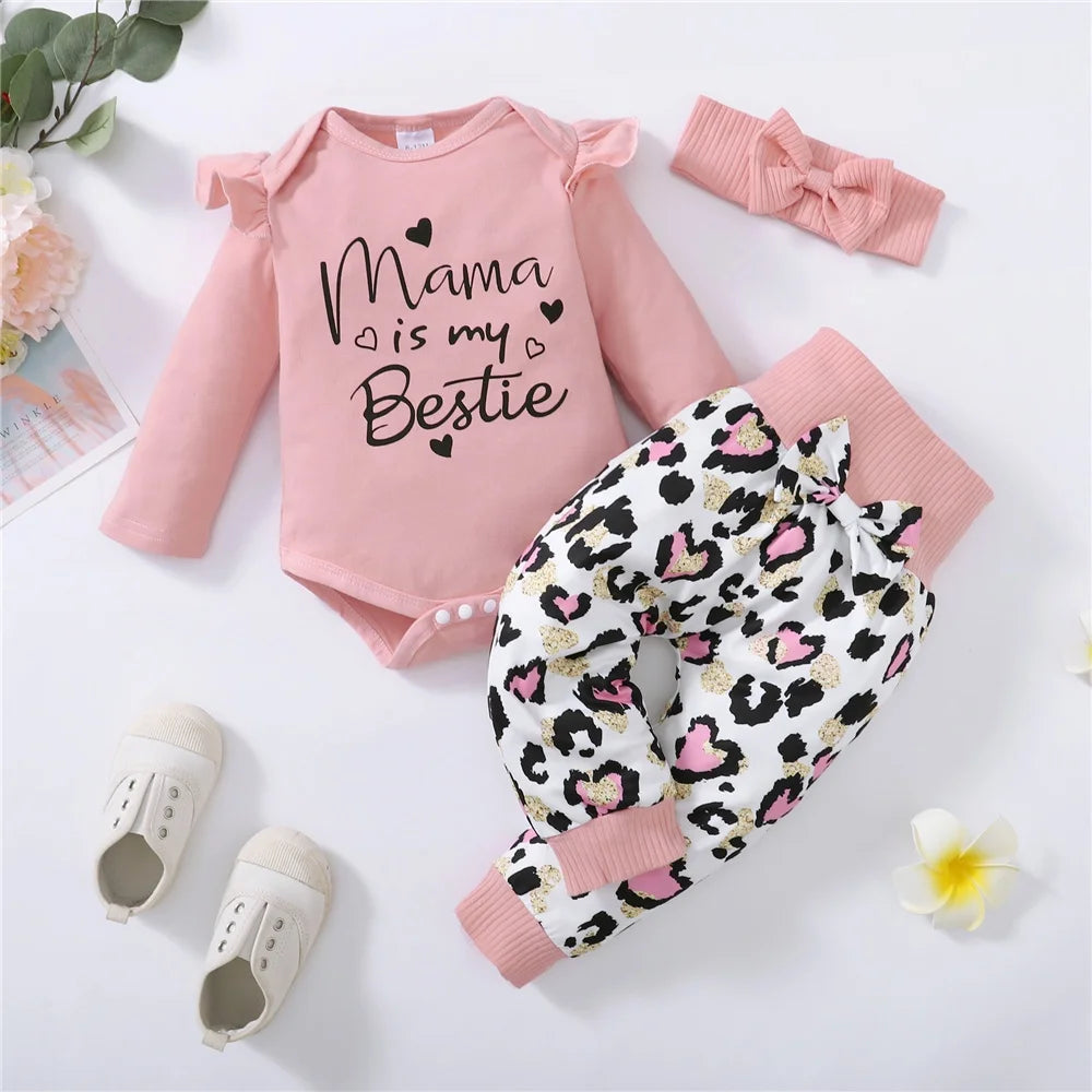 Pink baby outfit with "Mama is my Bestie" text, featuring a long-sleeve onesie, leopard print pants with pink cuffs, matching headband, cream shoes, and floral decor.