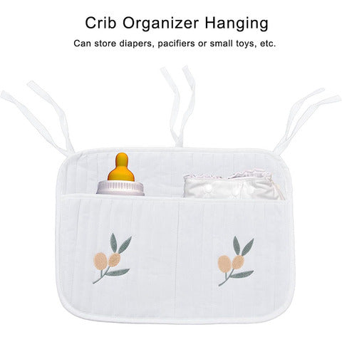 White crib organizer with two pockets, tied with strings. Holds a baby bottle and diapers, adorned with simple leaf and flower embroidery. Text above reads: "Crib Organizer Hanging."