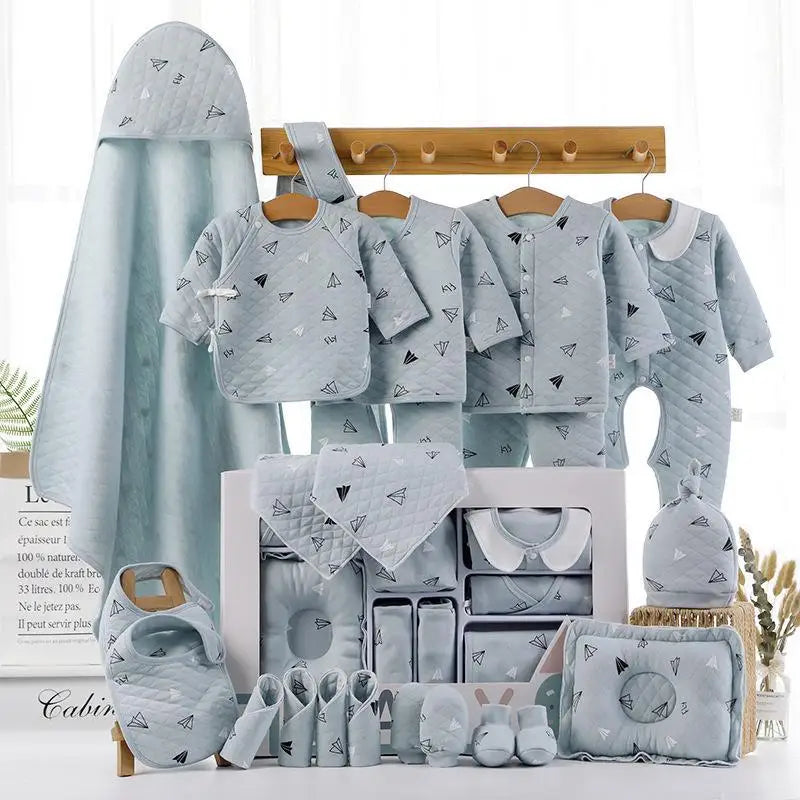 A collection of baby clothes and accessories in light blue with paper airplane patterns. Items include shirts, pants, a hat, bib, booties, mittens, and towels, displayed on wooden hooks.