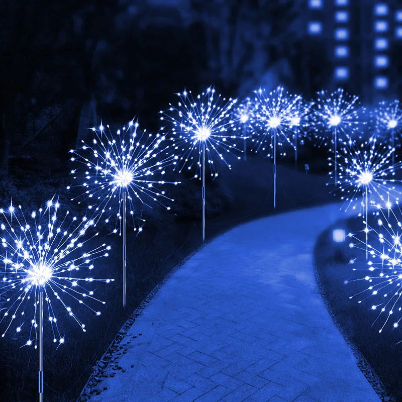 A winding path is lined with glowing blue LED dandelion lights in a dark park, creating a magical, illuminated scene.