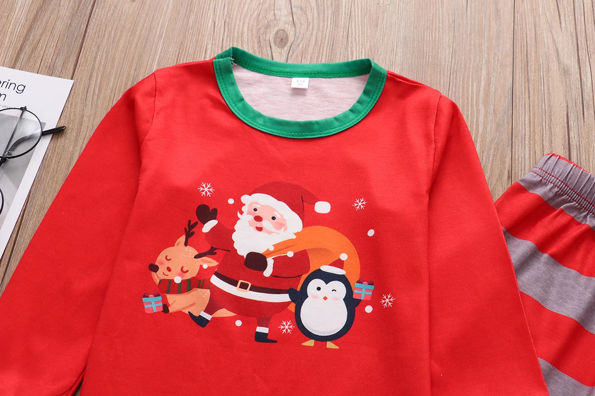 Red holiday-themed pajama top with a green collar featuring Santa, a reindeer with a scarf, and a penguin holding gifts. Glasses are beside the shirt on a wooden surface.