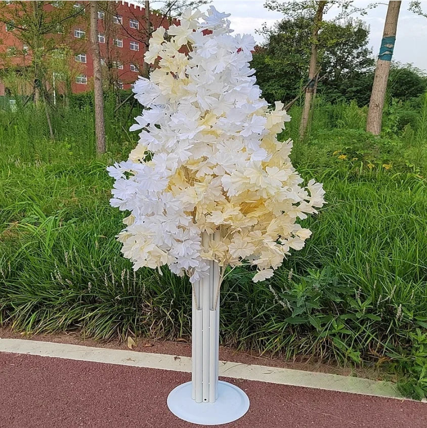 Artificial tree with white and cream leaves on a white stand, set against a grassy background with trees and a brick building partially visible.