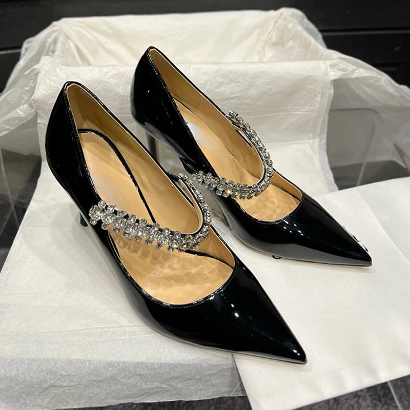 Elegant black patent leather high heels with pointed toes and sparkling rhinestone straps, displayed on white tissue paper inside a box.