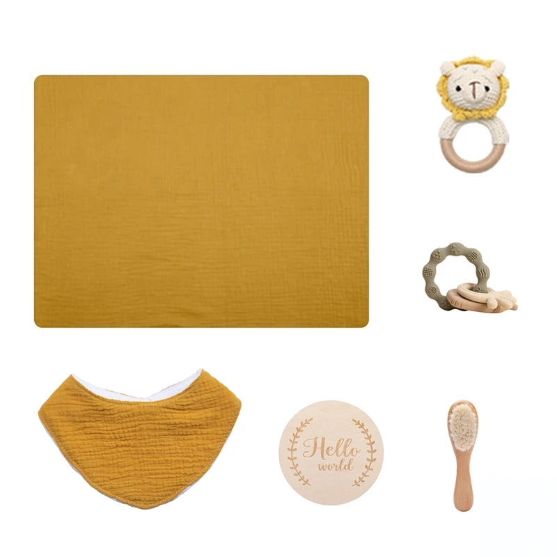 A baby gift set with a mustard muslin blanket, lion rattle, teether, bib, wooden "Hello World" plaque, and soft-bristle brush, all arranged on a white background.