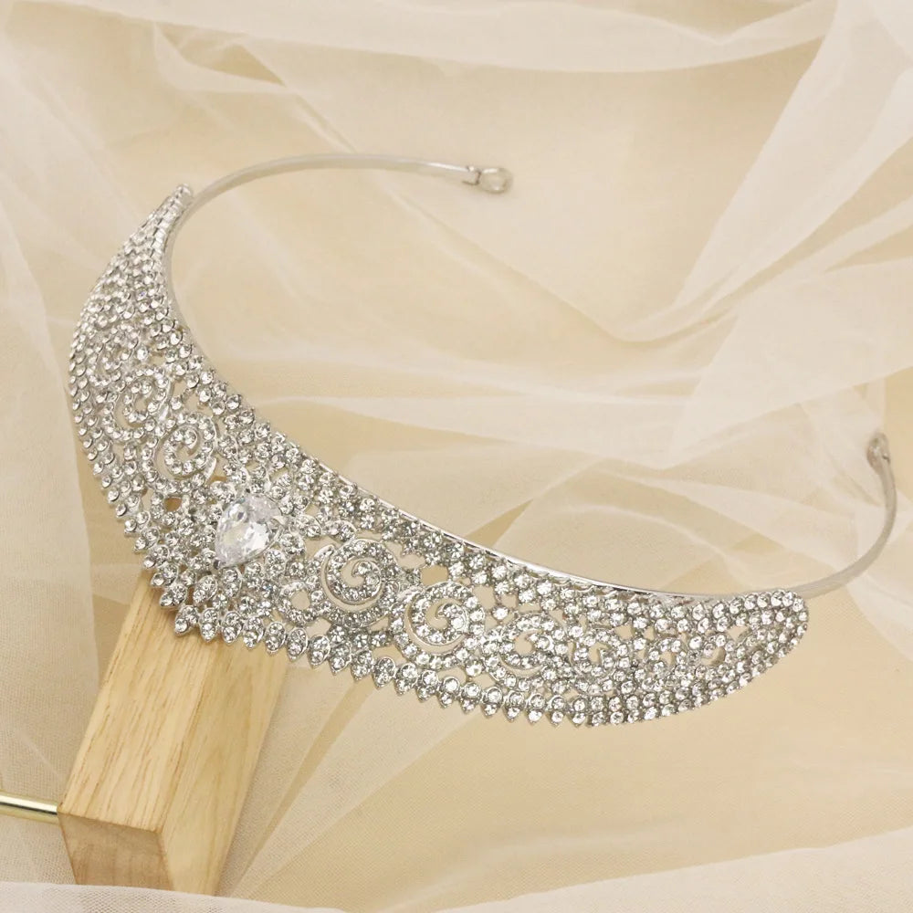 Elegant silver tiara encrusted with sparkling crystals and a central teardrop gem, resting on a wooden stand against a soft beige fabric background.