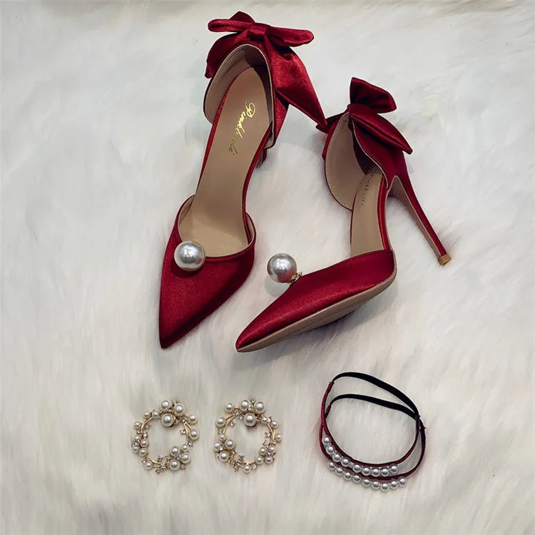 Red satin high heels with pearls and bows, displayed on white fur. Accompanied by pearl hoop earrings and two pearl bracelets.