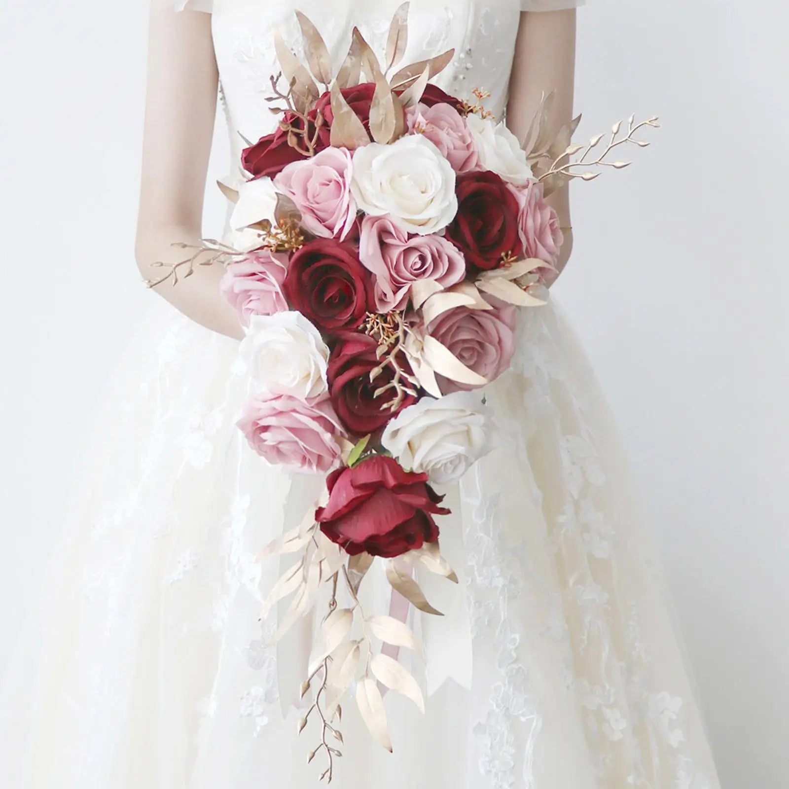 A bride in a lace dress holds a cascading bouquet of red, pink, and white roses, accented with gold leaves against a plain background.