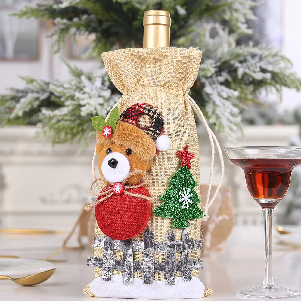 A gold wine bottle bag decorated with a plush bear, red fabric circle, green glittery tree, and wooden fence sits on a table. A glass of red liquid is beside it, with a snowy tree backdrop.
