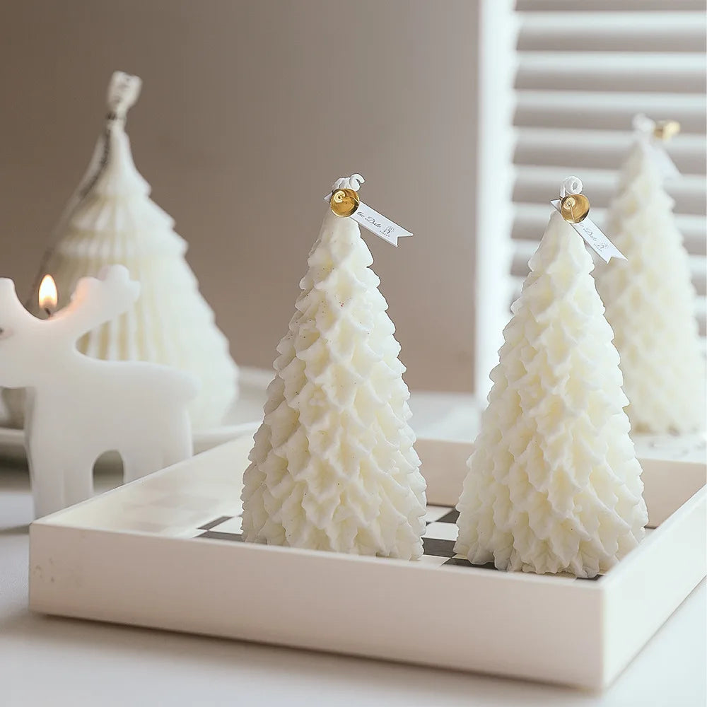 Decorative white candles shaped like Christmas trees are placed on a white tray. A small white reindeer candle holder with a lit candle is in the background.