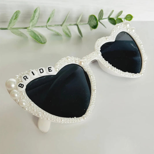 Heart-shaped sunglasses with pearl decoration and the word "BRIDE" in beads on the top left. A green leafy branch is in the background on a white surface.