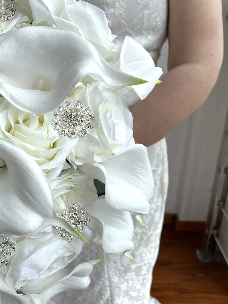 Bride holding a bouquet of white calla lilies and roses, adorned with sparkling rhinestone embellishments, against a lace wedding dress background.