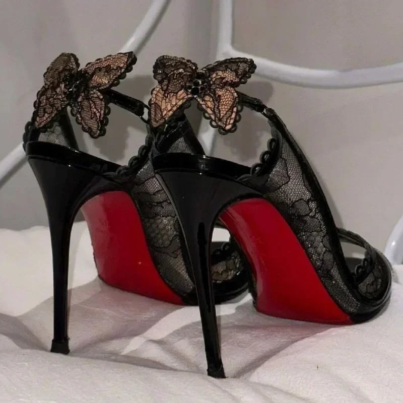 Black heels with lace detailing and butterfly accents, featuring iconic red soles. Stylishly poised on a white surface, highlighting their elegant design.