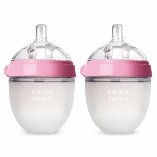 Two silicone baby bottles with pink lids and clear silicone nipples. The brand name "Comotomo" is printed on each bottle. The bottles are designed to be squeezable.
