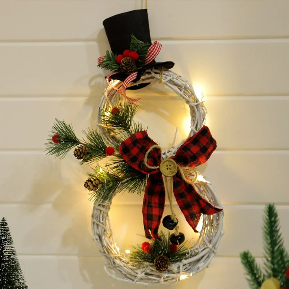 A festive wreath shaped like a snowman hangs on a wall, adorned with a black hat, plaid bow, pine branches, and lights, creating a cozy holiday decoration.