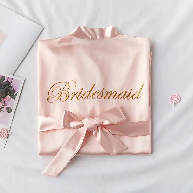 Folded pale pink satin robe with "Bridesmaid" embroidered in gold script. The robe is tied with a matching ribbon. A magazine and tiny lollipop are nearby on a white surface.