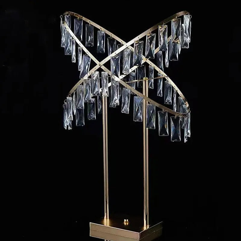 A modern decorative lamp with intersecting gold rings adorned with hanging rectangular crystals, mounted on a square base against a black background.
